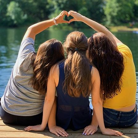 Trois jeunes femmes formant un cœur avec leurs bras au bord d’un lac, symbole d’une entreprise à impact engagée pour l’eau et la solidarité